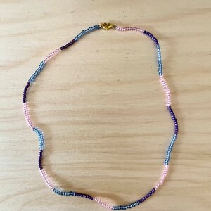 May include: A multi-colored beaded necklace with pink, blue, and purple beads. The necklace is made of small, round beads and has a gold clasp.