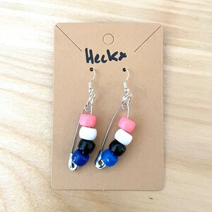 Könnte beinhalten: Ein Paar Ohrringe mit Sicherheitsnadeln und bunten Perlen. Die Perlen sind rosa, weiß, schwarz und blau. Die Ohrringe befinden sich auf einer braunen Karte mit dem Wort "Heckx" darauf.