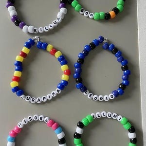 May include: Six colorful beaded bracelets with white letter beads spelling out names. The bracelets feature various color combinations, including purple, green, blue, pink, and black. The names include Killua, Gon, Kurapika, Leorio, Hisoka, and Illumi.