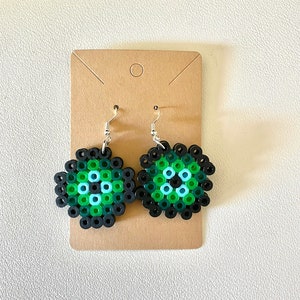 May include: A pair of green and black eye-shaped earrings made from perler beads. The earrings are hanging from silver hooks.