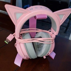 Puede incluir: Auriculares de juego de color rosa con orejas de gato y un soporte rosa. Los auriculares tienen una almohadilla gris y un cable USB.