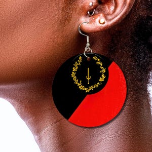 Puede incluir: Un par de pendientes grandes y redondos con un diseño negro y rojo. Los pendientes presentan una corona de laurel dorada con una espada en el centro.