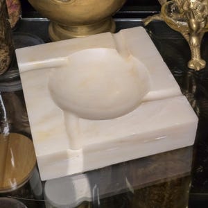 May include: A square, off-white marble ashtray with a circular indentation in the center. The ashtray has a smooth, polished surface and a rectangular shape. The ashtray is a decorative item.