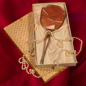Puede incluir: Un espejo de mano antiguo con marco y mango dorados, presentado en una caja decorativa. La superficie del espejo es de color rosa polvoriento y roto. Un collar de perlas falsas está drapeado alrededor del espejo y la caja, sobre un fondo rojo.