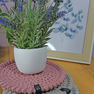 Op de afbeelding: Drie ronde gehaakte onderzetters in roze, beige en grijs. De onderzetters zijn op elkaar gestapeld. Een witte pot met een lavendelplant staat op de onderzetters.