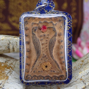 Puede incluir: Colgante rectangular con un diseño central marrón, que presenta una figura estilizada con alas y un acento floral dorado. Una gema roja se encuentra en la parte superior. El colgante está encerrado en un marco azul con detalles dorados.