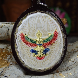 May include: A white and gold amulet with a red, green, and gold Garuda figure in the center. The amulet is surrounded by a white border with gold accents and is set in a dark brown oval frame.