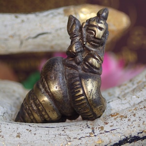 Puede incluir: Pequeña figura de latón de estilo antiguo que representa una deidad o figura mítica montada en una concha. La figura tiene rasgos faciales detallados y está adornada con intrincados detalles. La concha tiene un diseño en espiral.