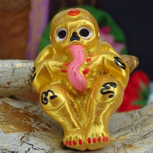 May include: A small golden figurine of a seated figure with a long pink tongue sticking out. The figure has black eyes and is decorated with black markings.