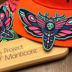May include: A pink and blue embroidered moth patch with a skull design. The patch is attached to a bright orange baseball cap. The cap is sitting on a wooden box with the text "Project Manticore" engraved on it.