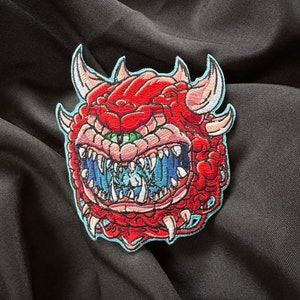 May include: A red and blue embroidered patch of a monster with sharp teeth and a large eye. The monster has a light blue outline.