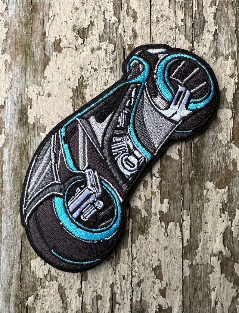 May include: An embroidered patch depicting a futuristic motorcycle with a single wheel, in black and blue.