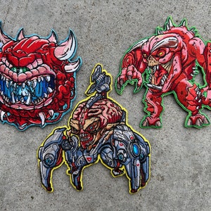 May include: Three embroidered patches featuring monsters from a video game. The patches depict a red demon with horns, a red monster with sharp teeth, and a robotic creature with a brain-like head. Each patch has a detailed design and colorful stitching.