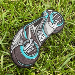 May include: An embroidered patch depicting a futuristic motorcycle in black and grey with a teal blue wheel. The patch is displayed on a green grassy background.