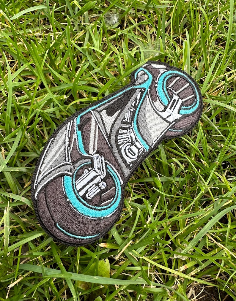 May include: An embroidered patch depicting a futuristic motorcycle in black and grey with a teal blue wheel. The patch is displayed on a green grassy background.