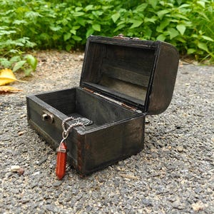 Könnte beinhalten: Eine kleine, rechteckige, dunkel gebeizte Holztruhe mit Klappdeckel, die offen einen leeren Innenraum zeigt. Eine silberfarbene Kette mit einem facettierten, sechseckigen, rotbraunen Steinanhänger hängt über der Vorderkante der Truhe.