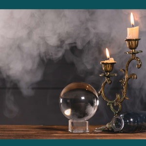 May include: A clear glass crystal ball sits on a wooden surface in front of a vintage brass candlestick with two lit candles. Smoke curls around the candles and the crystal ball.