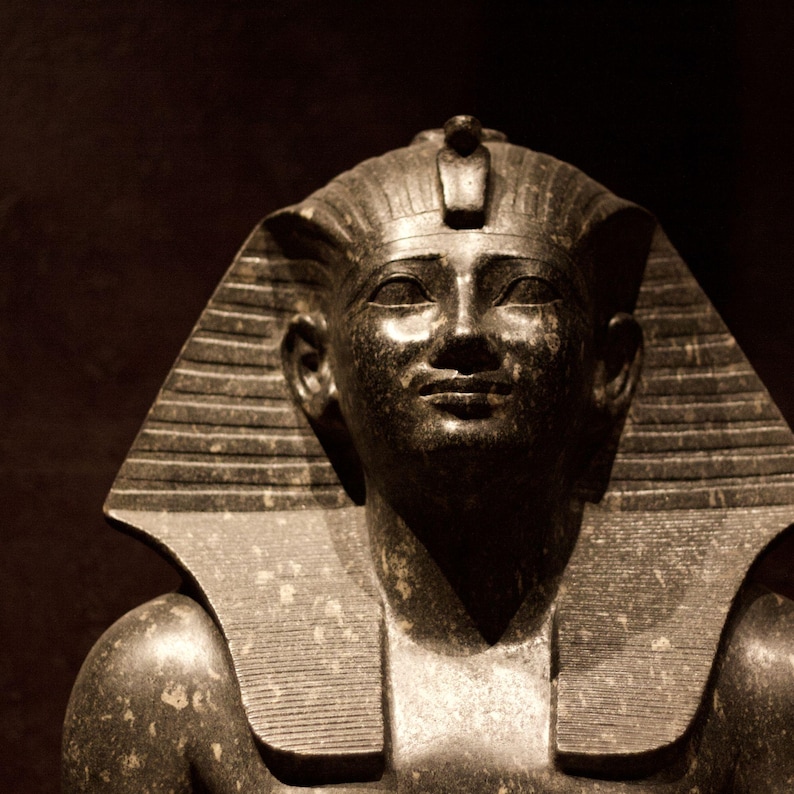 May include: A dark stone bust of an Egyptian pharaoh. The sculpture shows a serene face with detailed features, a striped headdress, and a ceremonial collar. The bust is set against a dark background, highlighting the intricate details.