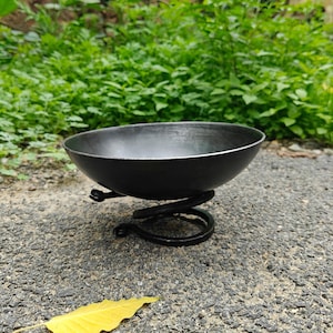 Puede incluir: Un cuenco circular de metal gris oscuro con un acabado liso y mate. El cuenco se apoya sobre una base construida a partir de una sola pieza de metal negro enrollada que forma un soporte en espiral, elevando el cuenco sobre la superficie.