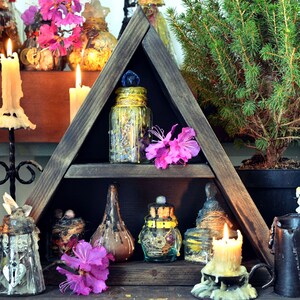 Pode incluir: Uma prateleira triangular de madeira com v&aacute;rias garrafas e frascos de vidro cheios de ervas e outros ingredientes. A prateleira &eacute; decorada com flores cor-de-rosa e velas.