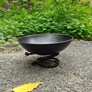 Puede incluir: Un cuenco circular de metal gris oscuro con un acabado liso y mate. El cuenco se apoya sobre una base construida a partir de una sola pieza de metal negro enrollada que forma un soporte en espiral, elevando el cuenco sobre la superficie.