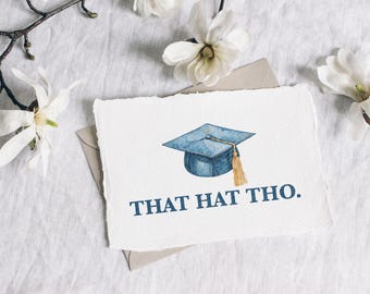 May include: A white card with a blue graduation cap illustration and the text "THAT HAT THO." The card is on a white linen background with white flowers.