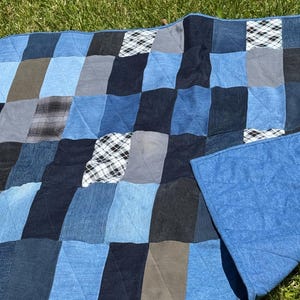 Op de afbeelding: Een patchwork quilt opgebouwd uit rechthoekige stukken stof in diverse tinten blauw, grijs en zwart denim. Het ontwerp bevat af en toe vierkanten met een zwart-wit ruitpatroon, met een achterkant van effen blauwe stof.
