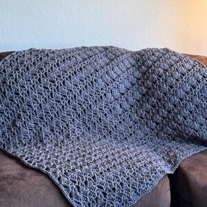 Puede incluir: Una manta gris tejida a crochet con un patrón calado texturizado, colocada sobre un sofá marrón. La manta muestra un diseño detallado y una textura suave. El color gris añade calidez y confort a cualquier espacio. Un elemento decorativo.