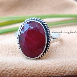 Könnte beinhalten: Ein silberner Ring mit einem roten ovalen Edelstein, der in eine gedrehte Silberband gefasst ist. Der Ring liegt auf einem braunen Stoffhintergrund.