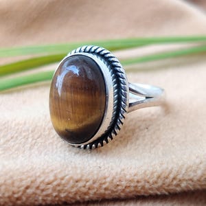 Puede incluir: Un anillo de plata con una piedra ovalada de ojo de tigre. El anillo tiene una banda decorativa con forma de cuerda.