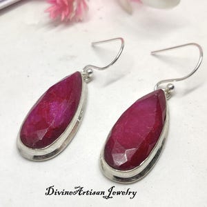 Könnte beinhalten: Ein Paar tropfenförmige Ohrringe mit facettierten, tiefroten Edelsteinen, die in silberfarbenem Metall gefasst sind. Die Ohrringe haben ein einfaches Haken-Design. Der Text "Divine Artisan Jewelry" befindet sich unten.