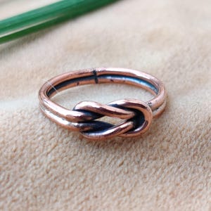 Könnte beinhalten: Ein Kupferring mit einem Knotendesign. Der Ring besteht aus zwei Kupferbändern, die miteinander verdreht sind, um einen Knoten zu bilden.
