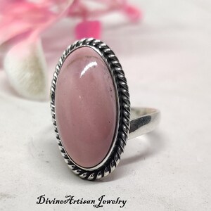May include: A silver ring with an oval, pink gemstone. The gemstone is set in a twisted rope-like border. The ring is on a white surface with pink flowers in the background. The text "Divine Artisan Jewelry" is at the bottom.