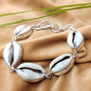 Könnte beinhalten: Ein silbernes Armband mit sechs weißen Kaurischnecken. Die Muscheln sind in silbernen Fassungen gefasst und durch silberne Glieder verbunden. Das Armband wird auf einem beigefarbenen Stoff mit grünen Blättern im Hintergrund präsentiert.