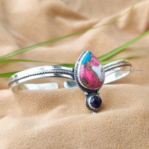 Può includere: Un bracciale a polsino in argento con una pietra a forma di goccia nei toni del rosa, bianco e turchese. Sotto la goccia c'è una piccola pietra rotonda viola. Il bracciale ha un design a corda intrecciata sui bordi.