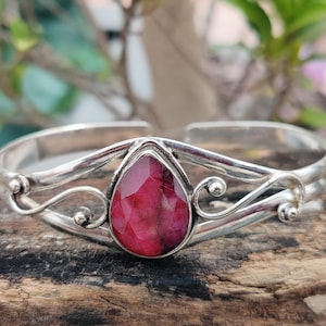Könnte beinhalten: Silbernes Armreif-Armband mit einem großen, tropfenförmigen, tiefroten Edelstein. Das Armband hat verzierte, wirbelnde Silberdesigns auf beiden Seiten des Edelsteins, vor einem natürlichen Holzuntergrund.