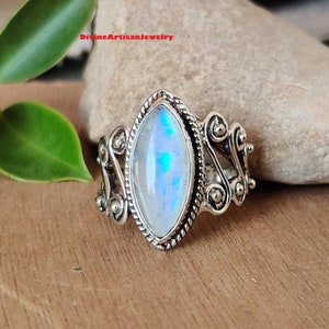 Könnte beinhalten: Ein silberner Ring mit einem großen, schillernden Mondstein, der in einem detaillierten, verzierten Design gefasst ist. Der Ring liegt auf einer Holzoberfläche.