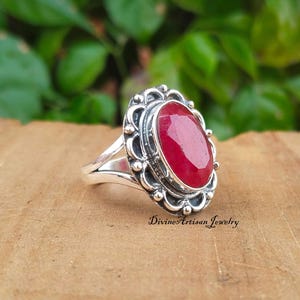 Könnte beinhalten: Ein silberner Ring mit einem großen, ovalen, roten Edelstein, der in einer detaillierten, verzierten Silberfassung gefasst ist. Der Ring liegt auf einer Holzoberfläche.