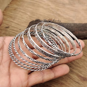 May include: A set of eight silver bangles with different textures and designs. The bangles are stacked on top of each other and have a shiny finish.