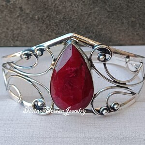 Könnte beinhalten: Ein silbernes Armreif-Armband mit einem großen, tropfenförmigen roten Edelstein, der in der Mitte eingesetzt ist. Das Armband hat ein zartes, verziertes Design mit Schwüngen und Kurven.