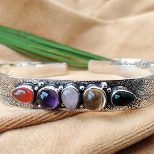 May include: Silver cuff bracelet with an engraved floral pattern. The bracelet features five colourful gemstones: red, purple, pink, yellow-brown, and black. The text "Divino Artisan Jewelry" is visible on the bracelet.