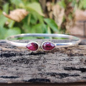 Könnte beinhalten: Ein silbernes Armband mit zwei tropfenförmigen, tiefroten Edelsteinen. Das Armband hat ein schlichtes Design mit den Edelsteinen an der Vorderseite. Der Hintergrund ist eine Holzoberfläche mit grünem Laub.