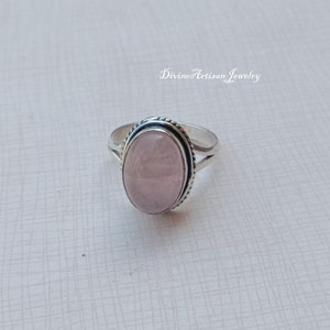 Könnte beinhalten: Ein silberner Ring mit einem hellrosa ovalen Edelstein, der in einer Zargenfassung gefasst ist. Der Ring hat ein zartes, gedrehtes Band.