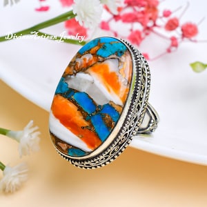 Könnte beinhalten: Ein silberner Ring mit einem ovalen Türkis-, Orange- und Weißstein. Der Ring hat ein dekoratives silbernes Band.