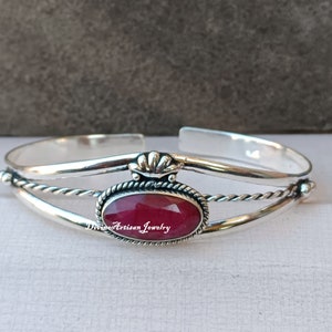 Könnte beinhalten: Ein silbernes Armreif-Armband mit einem gedrehten Seil-Design und einem roten Edelstein, der in einer ovalen Silberfassung sitzt. Das Armband hat auf einer Seite eine dekorative silberne Krone.