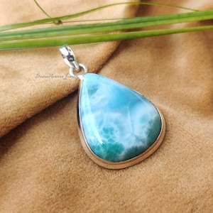 May include: A teardrop-shaped pendant with a light blue and white stone set in a silver-colored metal frame. The stone has a marbled appearance with shades of turquoise and white. The pendant is hanging from a silver-colored bail.