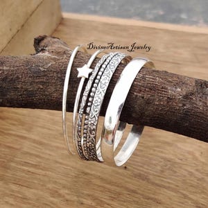 Könnte beinhalten: Satz von fünf silbernen Armreifen, einer mit einem Sternanhänger, einer mit einem Perlmuster und drei mit Blumenmustern. Die Armreifen sind auf einem Holzast gestapelt. Divine Artisan Jewelry.