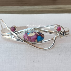 Könnte beinhalten: Silberarmband mit einem rosafarbenen und blauen Edelstein sowie zwei kleineren rosafarbenen Edelsteinen. Das Armband hat ein zartes, fließendes Design.