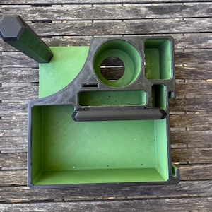 Peut inclure: Un organiseur de bureau imprimé en 3D, noir et vert, avec plusieurs compartiments. Il comprend un grand support circulaire, un plateau rectangulaire et plusieurs petites fentes. L'organisateur est posé sur une surface en bois.