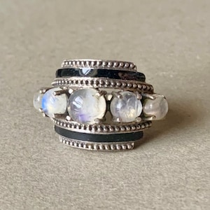 May include: A silver ring with four white moonstone gemstones set in a black enamel band.